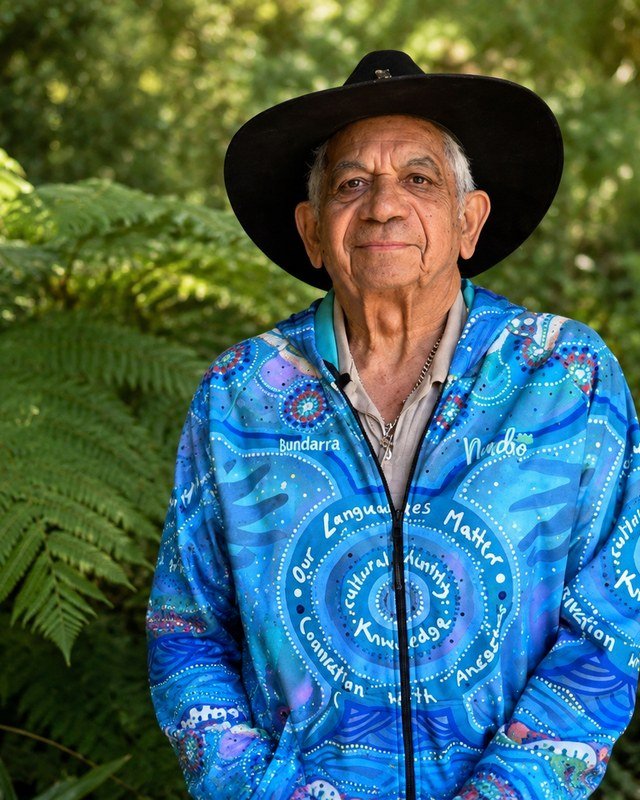 Uncle Bill Lemson standing among ferns in his black hat, wearing a blue shirt painted with the words Our Languages Matter.
