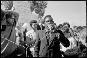 Dexter Daniels speaks into a microphone at an outdoor rally, with placards and a crowd of listeners around him.