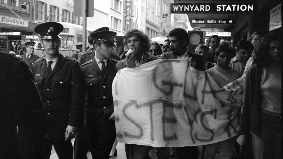 Marchers in a Sydney street carry a painted banner reading Gurindji and Vesteys past Wynyard Station, with uniformed police walking alongside them.