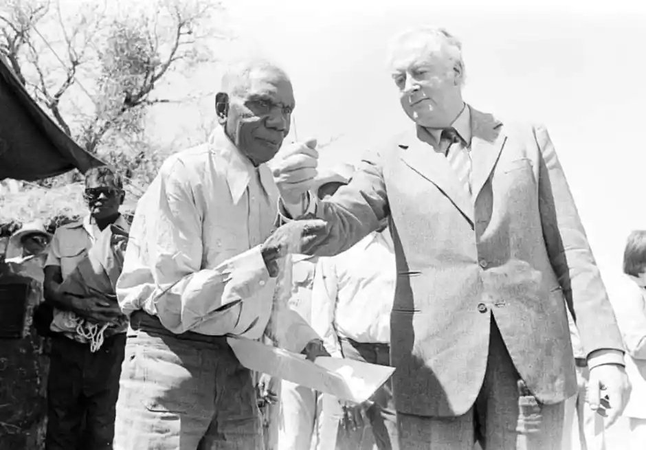 Vincent Lingiari holds documents in one hand and reaches out the other as Gough Whitlam pours soil into it, with Gurindji people standing behind them.