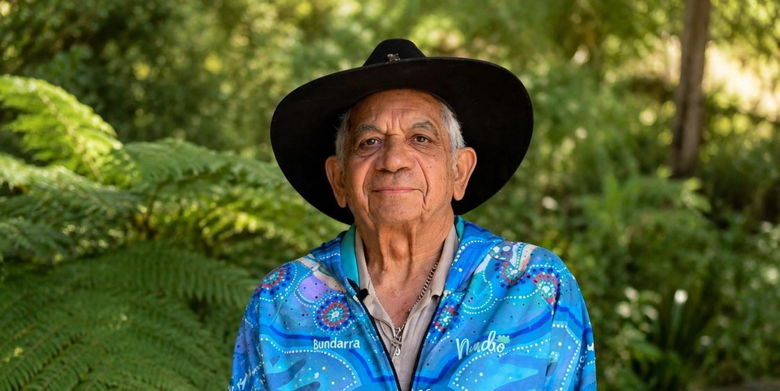 Uncle Bill Lemson, in his hat and a blue shirt with First Nations designs, standing among green tree ferns and looking toward the viewer
