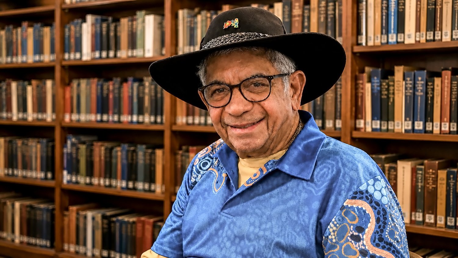 Uncle Bill Lemson smiling warmly in his black hat and blue shirt, shelves of books behind him