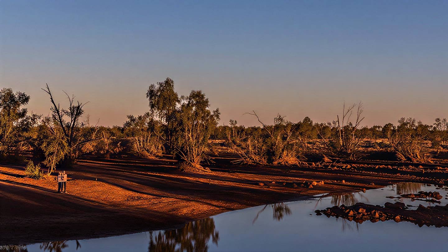 Burke River at evening, near Boulia, Central Western Queensland. Coolibah and river red gum on the bank, red iron-rich soil in the foreground catching the last light, water reflecting the dusk sky.