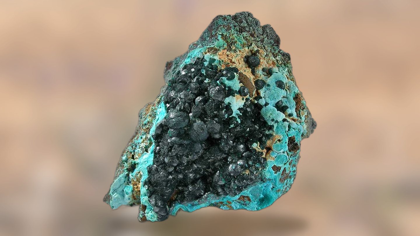 A piece of copper-bearing rock from the Cloncurry District, turquoise chrysocolla crusts and dark green pseudomalachite over earthy host stone, resting on pale straw-coloured ground.