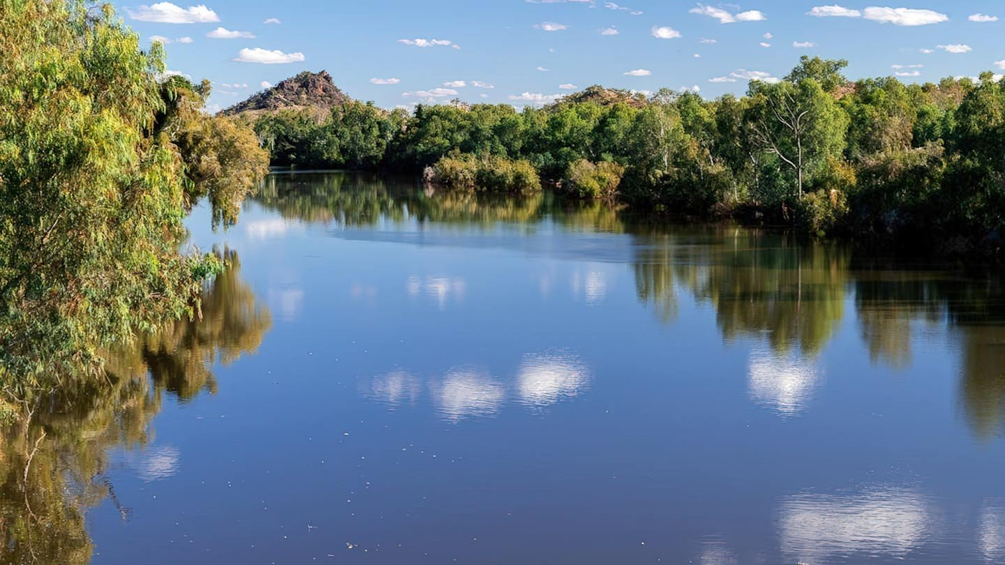 The Cloncurry River in north-west Queensland, with eucalypts on its banks and a granite outcrop in the middle distance. Still water carrying a deep slate-indigo, with sky and trees reflected on its surface.