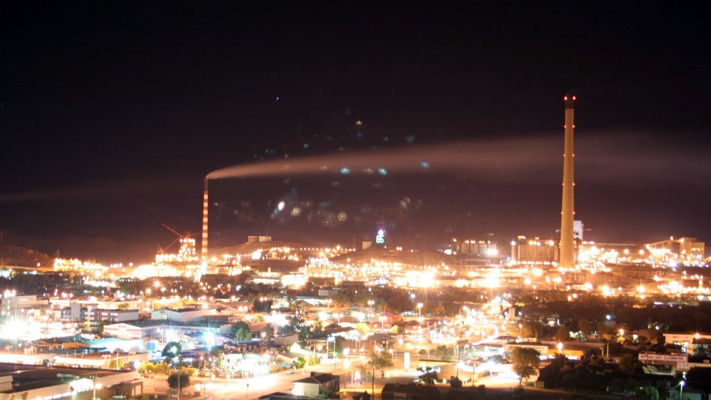 Mount Isa at night: the mine and town burning bright beneath a deep brown-black sky, the smelter stacks silhouetted against the dark.