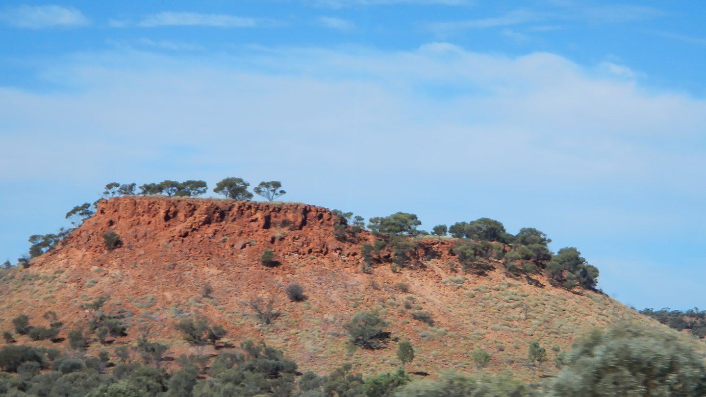 A weathered red-rock outcrop in Channel Country near Middleton, central western Queensland, with eucalypts on top and scrub at the base. The exposed iron-rich rock face has softened to a pale terracotta-orange under sun and time.