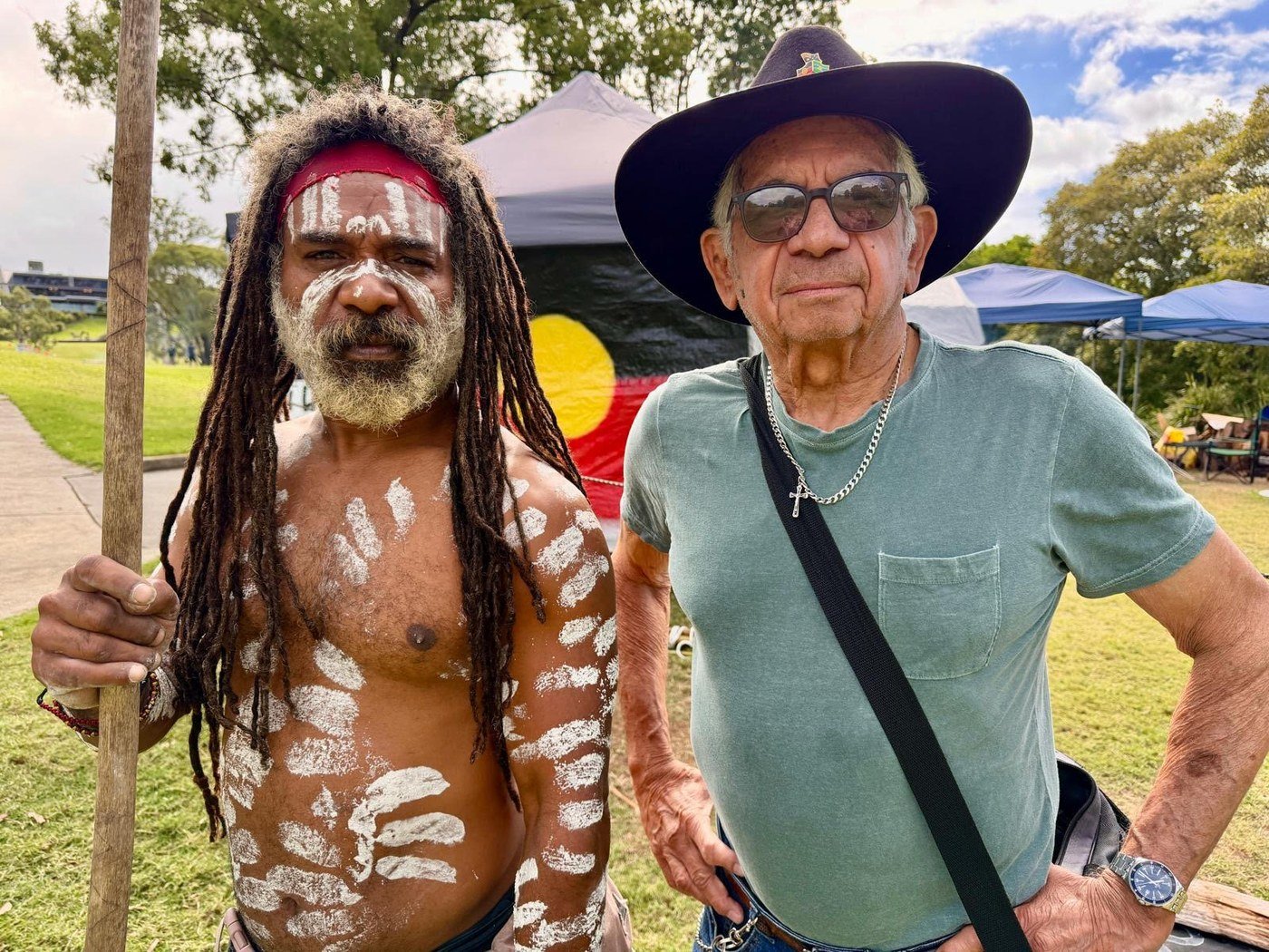 Uncle Derek &lsquo;Powder&rsquo; Oram (left) in cultural attire and ochre, standing with Uncle Bill Lemson (right) at the Goori Camp Embassy, with the Aboriginal flag in the background.