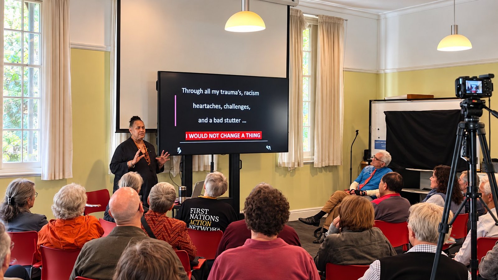 Aunty Sandra King OAM speaking to a seated audience in the Darnell Room, beside a screen that reads: Through all my trauma's, racism, heartaches, challenges, and a bad stutter, I would not change a thing.