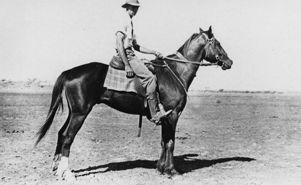 A horseman seated on a dark horse standing side-on in bare open country near Cloncurry, casting a hard shadow on the ground, with an empty horizon behind.