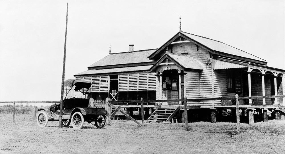 The post office at Burketown in 1920. A timber building raised on stumps with a corrugated iron roof, a gabled entrance porch reached by wooden steps, and a deep verandah running along the front. An early motor car with a folded canvas hood stands on the bare ground at the left, beside a telegraph pole.