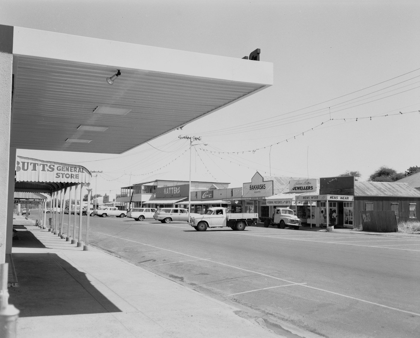 Ramsay Street, Cloncurry, in February 1966. Shopfronts along a wide street under deep awnings, with utes and station wagons parked at the kerb and strings of festive lights overhead. The shop signs read Butts General Store, Katters, Bakhashs, Jewellers and Mens Wear.
