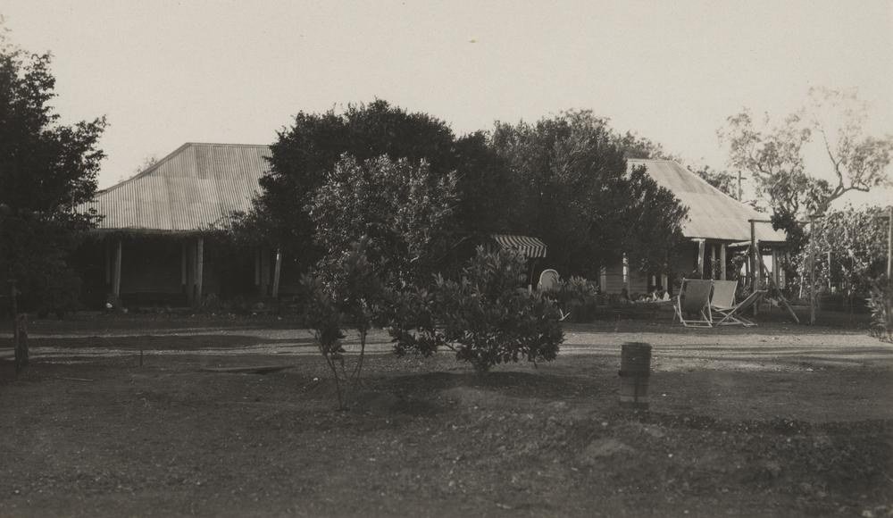 A low homestead on a station property in outback Queensland around 1925. The building has a corrugated iron roof and a deep shaded verandah, with a striped awning to one side, deck chairs standing on swept bare ground, and a young tree planted in the foreground.