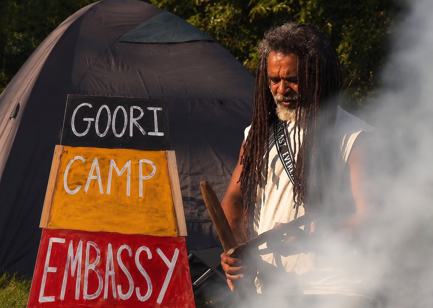 Uncle Derek &lsquo;Powder&rsquo; Oram conducting a smoking ceremony beside the Goori Camp Embassy sign (black, yellow and red panels lettered GOORI / CAMP / EMBASSY) at Barrambin, Brisbane.