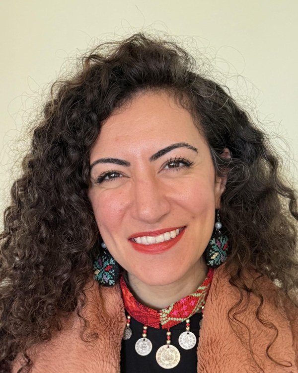 Remah Naji, a woman with long dark curly hair, smiling warmly, wearing embroidered Palestinian tatreez earrings and a traditional coin necklace.