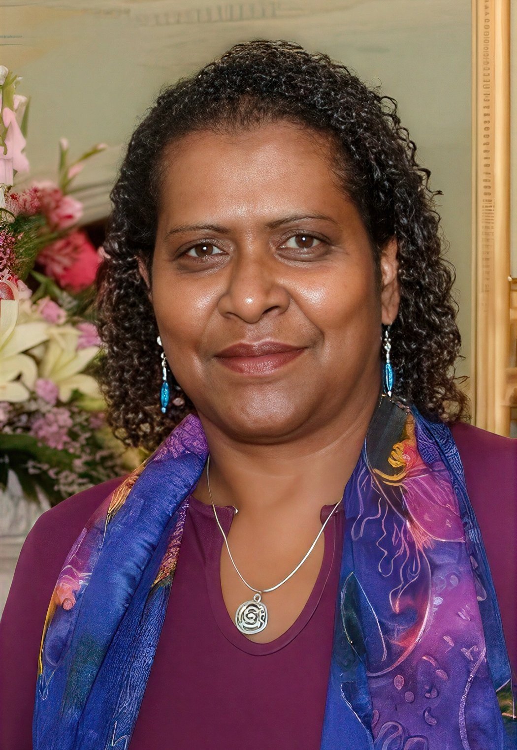 Dr Noritta Morseu-Diop, a First Nations woman with dark curly hair, wearing a deep purple top, a blue and purple silk scarf, and a silver pendant, smiling at the camera.